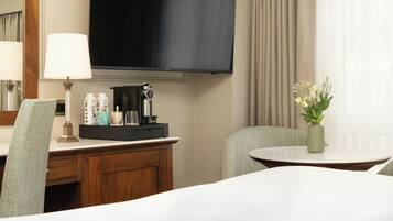Quarto de hotel com televisão de tela plana fixada acima de uma mesa de madeira, uma mesinha com um vaso de flores e uma cama com roupa de cama branca.