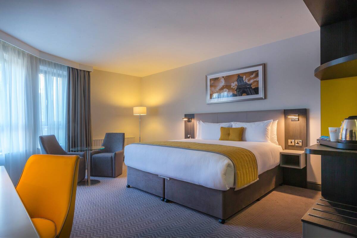 A hotel room with a bed, a desk, a chair, a lamp, and a painting on the wall.