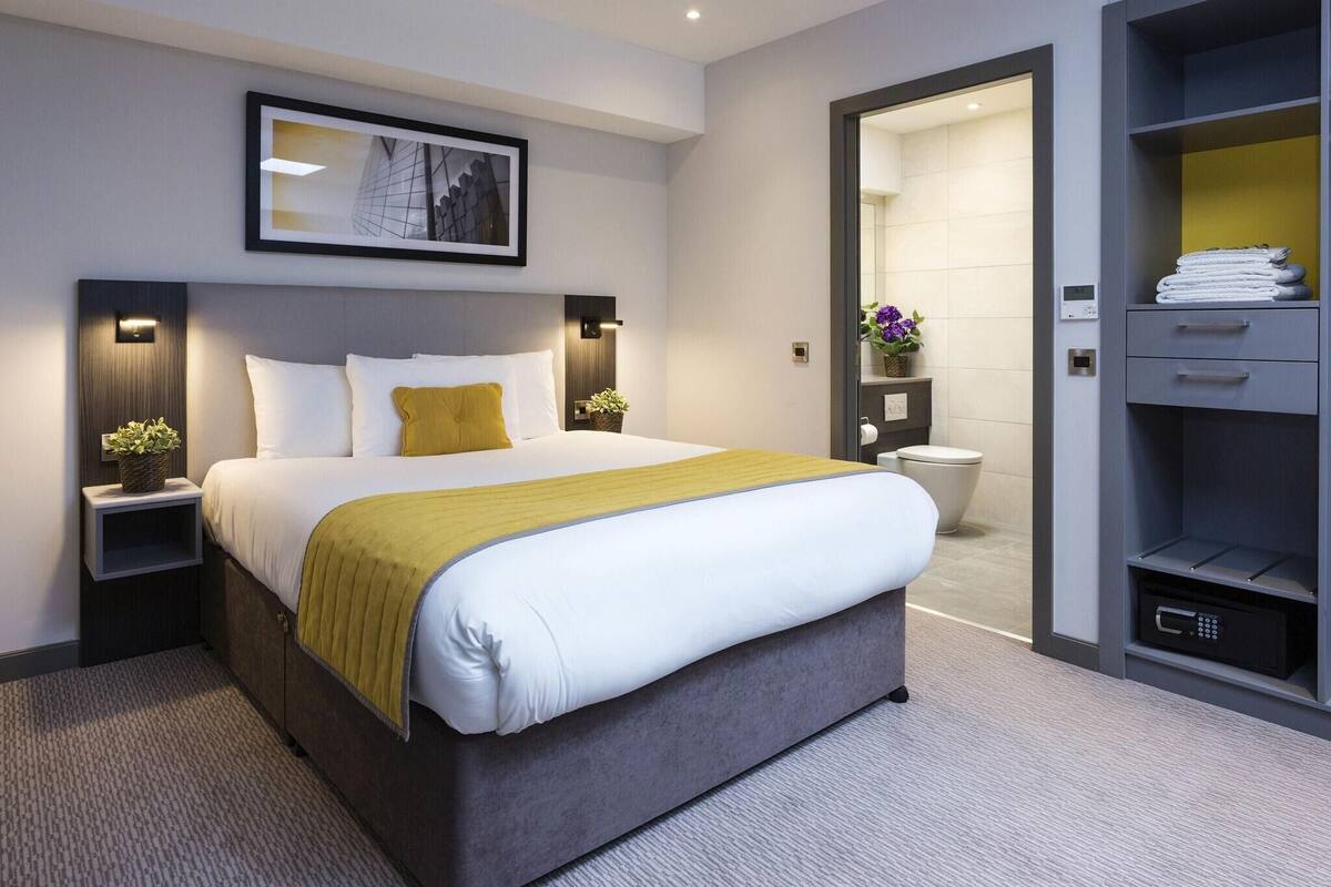 A modern hotel room with a large bed, bedside tables with lamps, a built-in wardrobe, and a bathroom visible through an open door.