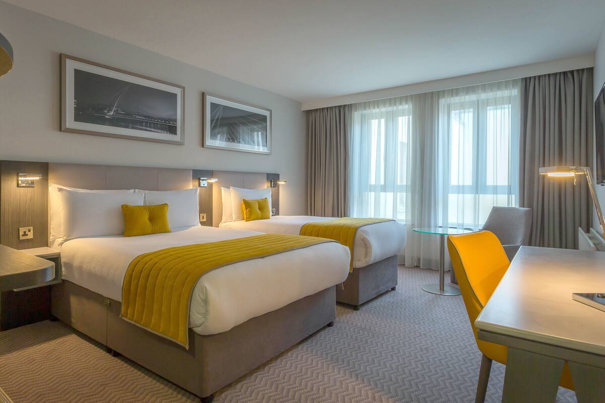 A hotel room with two beds, a desk, and a chair. There are two framed pictures on the wall, a coffee machine, and a lamp.