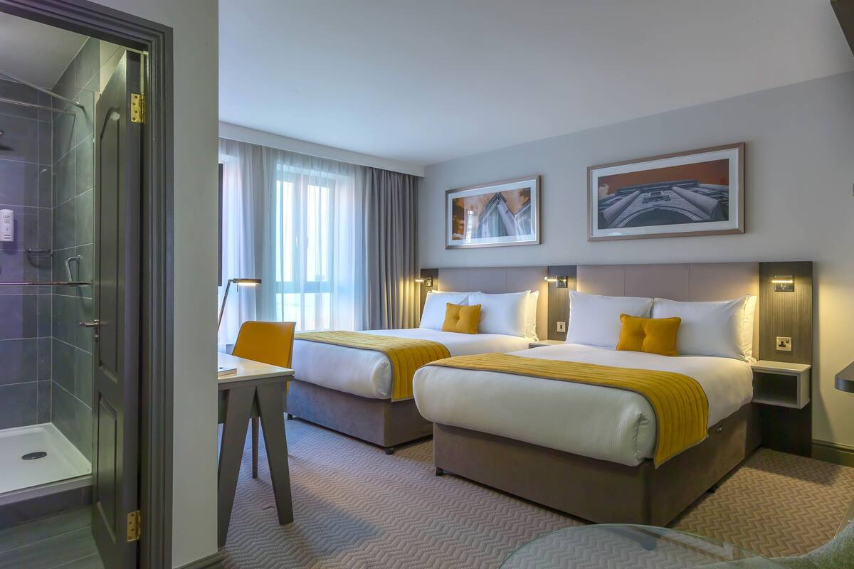 A hotel room with two beds, a desk, a chair, a coffee machine, and a shower area.