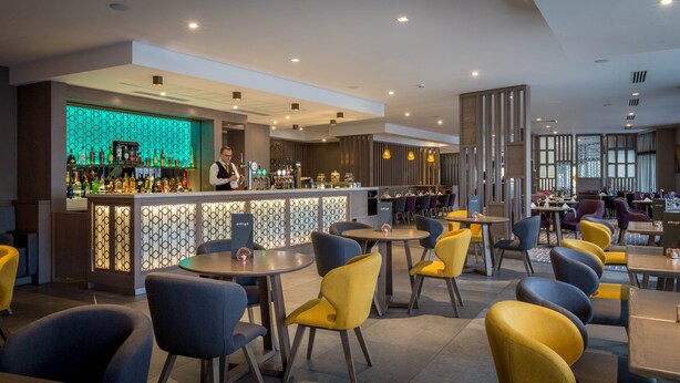 Un restaurante moderno con zona de bar, diversas opciones de asientos como mesas redondas y sillas, y una lámpara decorativa.