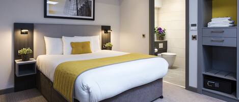 Una habitación de hotel moderna con una cama grande, mesitas de noche con lámparas, una planta junto a la cama, un cuadro en la pared, un vestidor con estantes y un baño visible a través de una puerta abierta.