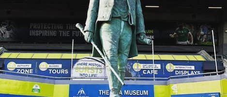 Una estatua de un hombre traje, sosteniendo un bate de béisbol, sobre un pedestal con el nombre 'CUSACK' grabado, frente a un edificio con varios anuncios.