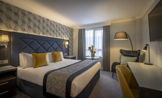 Habitación de hotel con una cama grande, una mesita de noche con lámpara, una silla, una pequeña mesa con un jarrón de flores y un televisor montado en la pared.