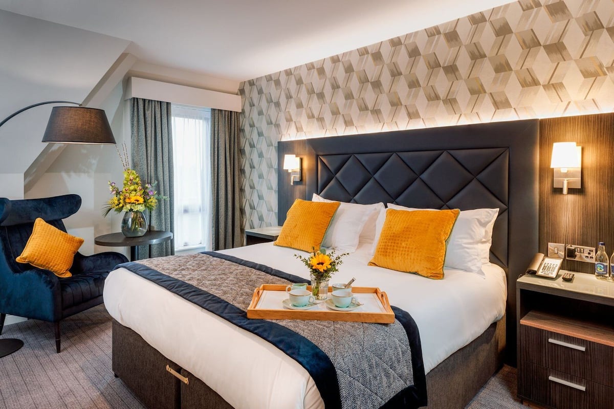 A hotel room with a large bed, a bedside table with a lamp, a chair, and a tray with cups and a vase of flowers.