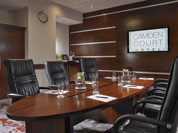 Uma sala de conferências com uma mesa comprida, cadeiras de couro preto, um relógio e uma placa com a inscrição 'Camden Court Hotel'.