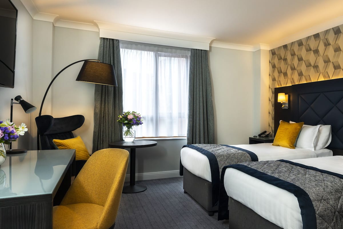 A hotel room with two beds, a desk, a chair, a lamp, and a window with curtains.