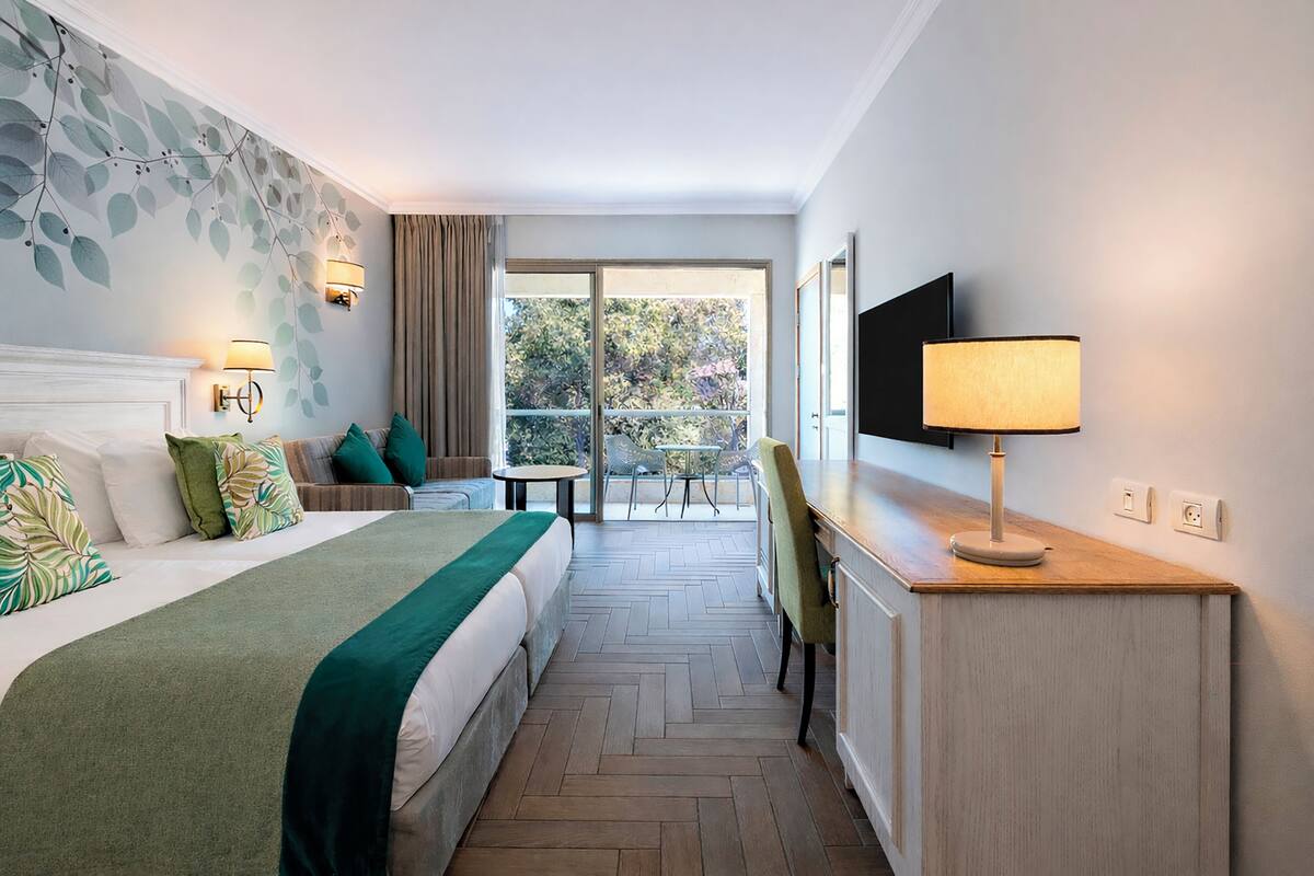 Ein Hotelzimmer mit Bett, Schreibtisch mit Lampe, Fernseher, Balkon mit Tisch und Stühlen sowie Blick auf Bäume.