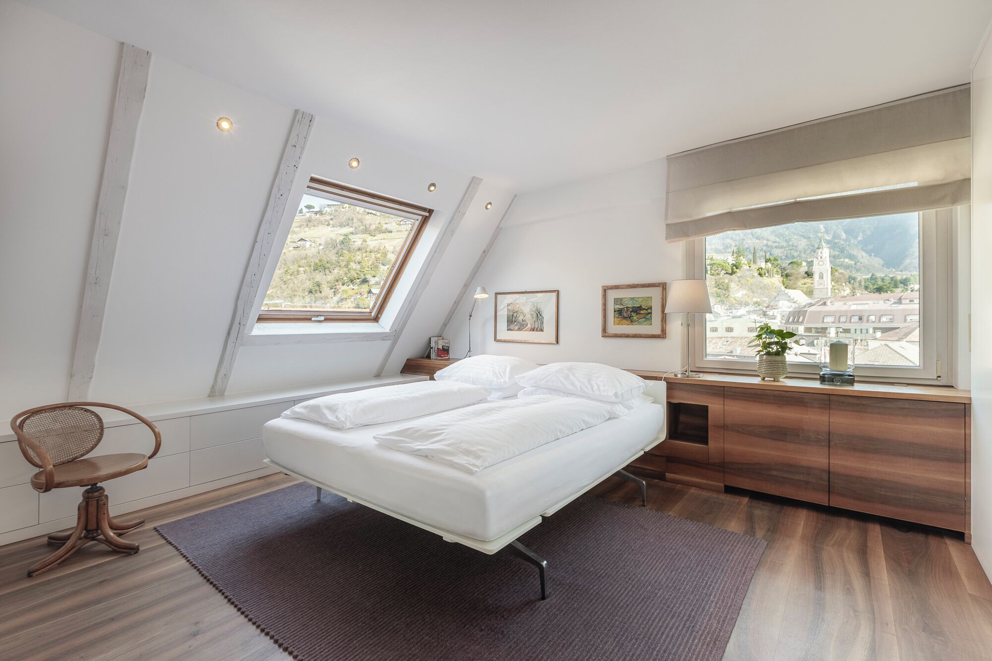 Een slaapkamer met een bed, een stoel, een bureau en een raam met uitzicht op gebouwen en bomen.