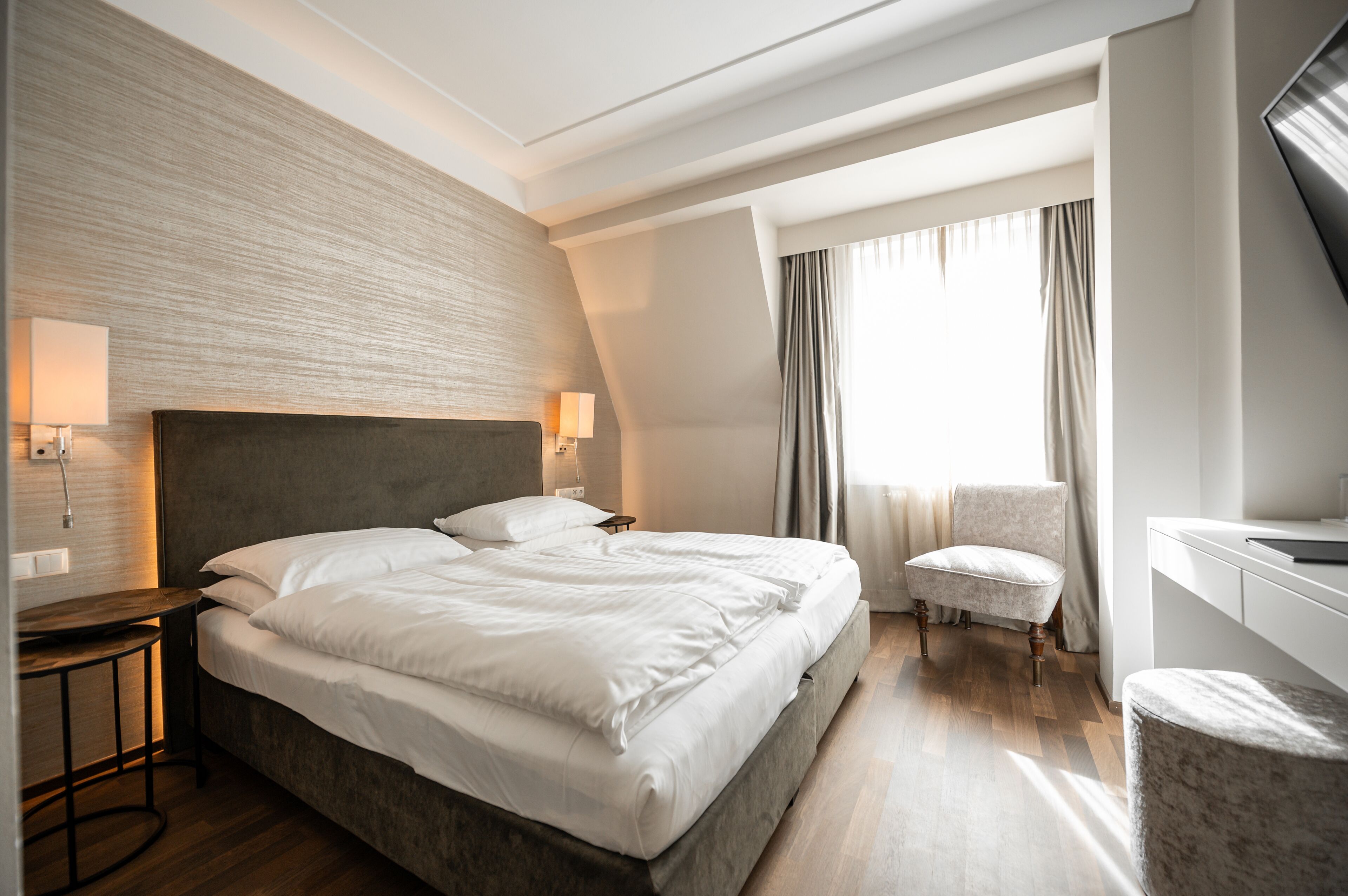 A modern hotel room with a large bed, a desk with a lamp, a chair, and a view of the outdoors.