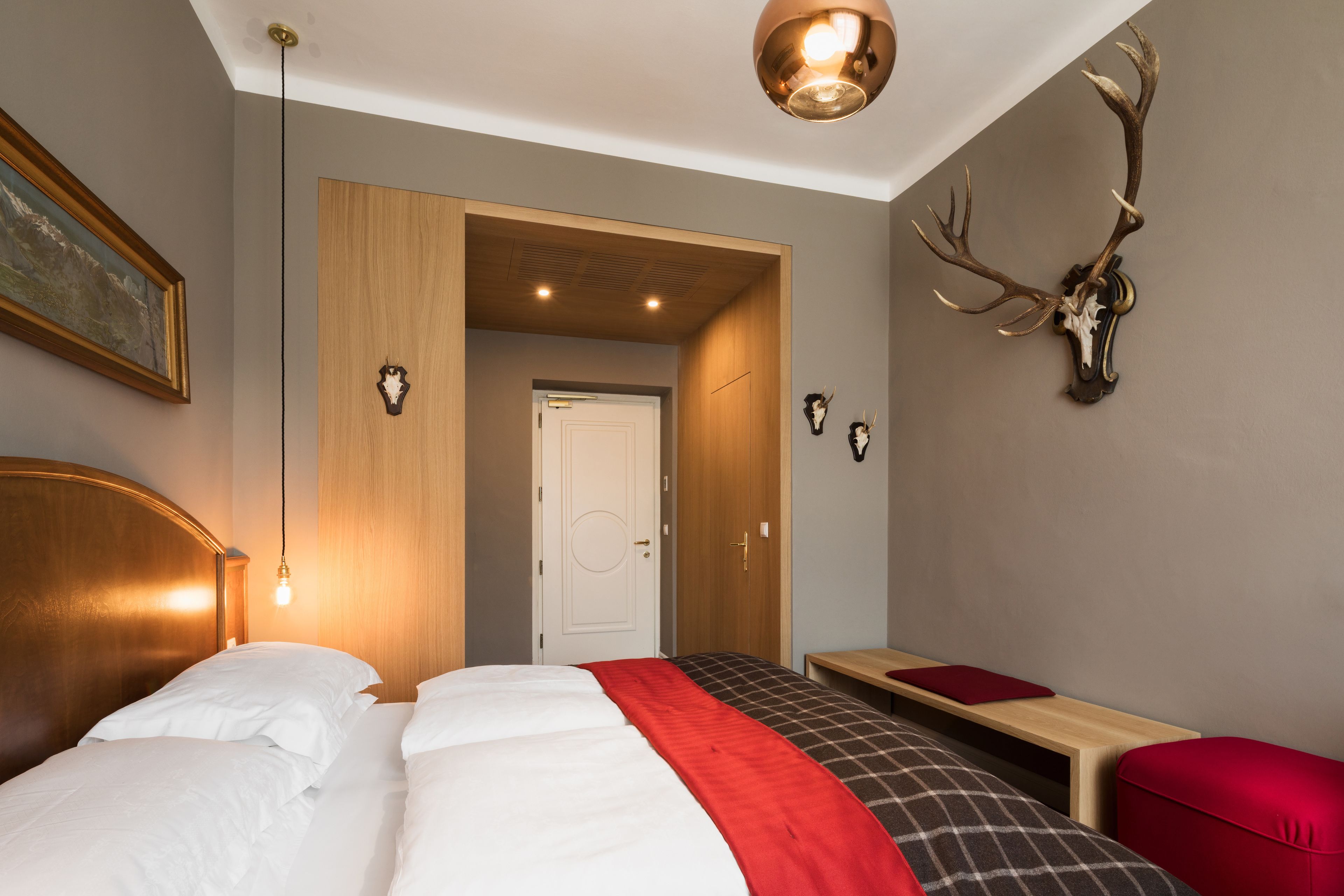 Una habitación de hotel con una cama grande, un cabecero de madera, una colcha a cuadros roja y negra, un banco de madera y una cabeza de venado montada en la pared.