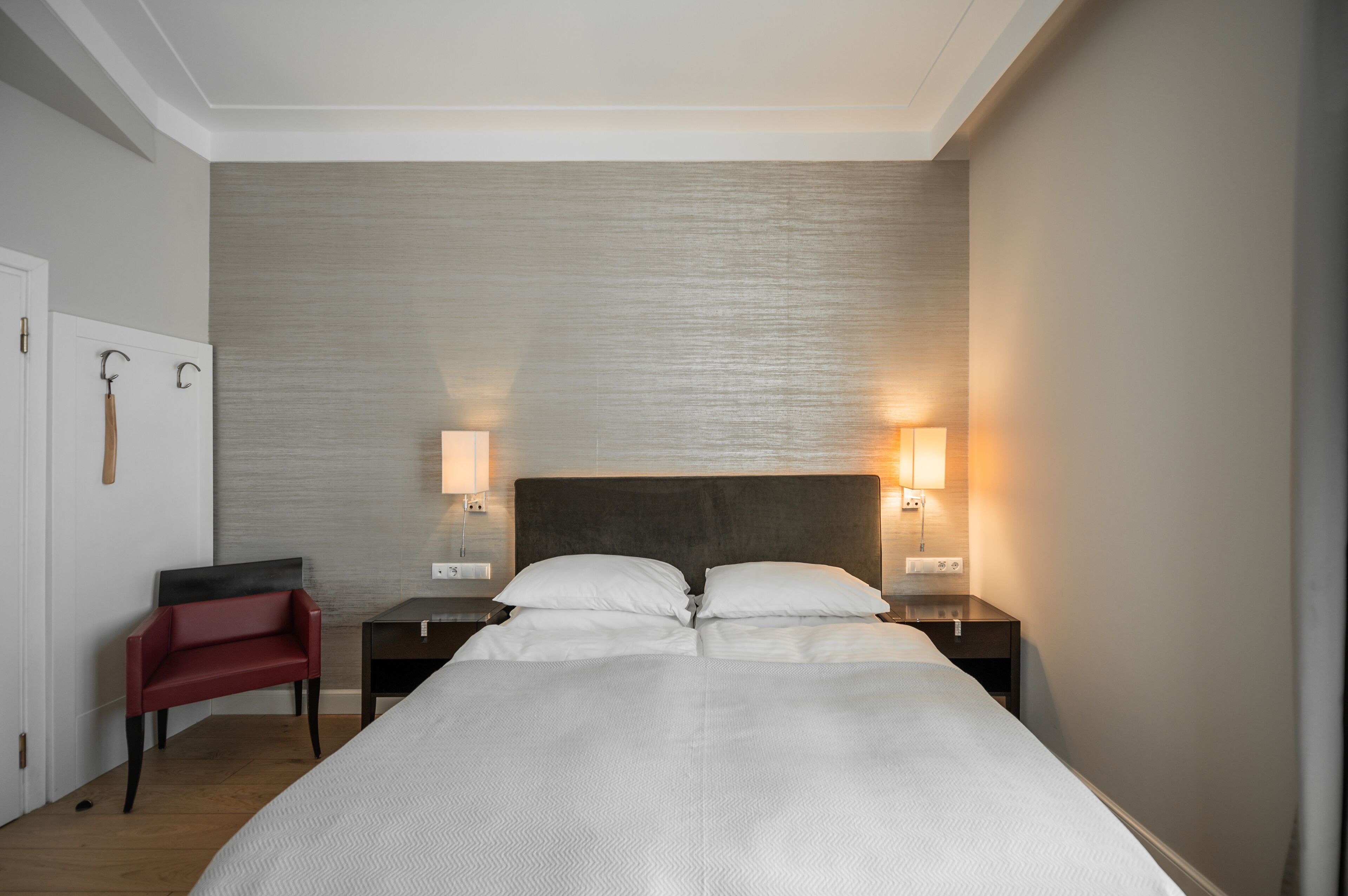 A hotel room with a large bed, two bedside tables with lamps, a red chair, and a wooden door.
