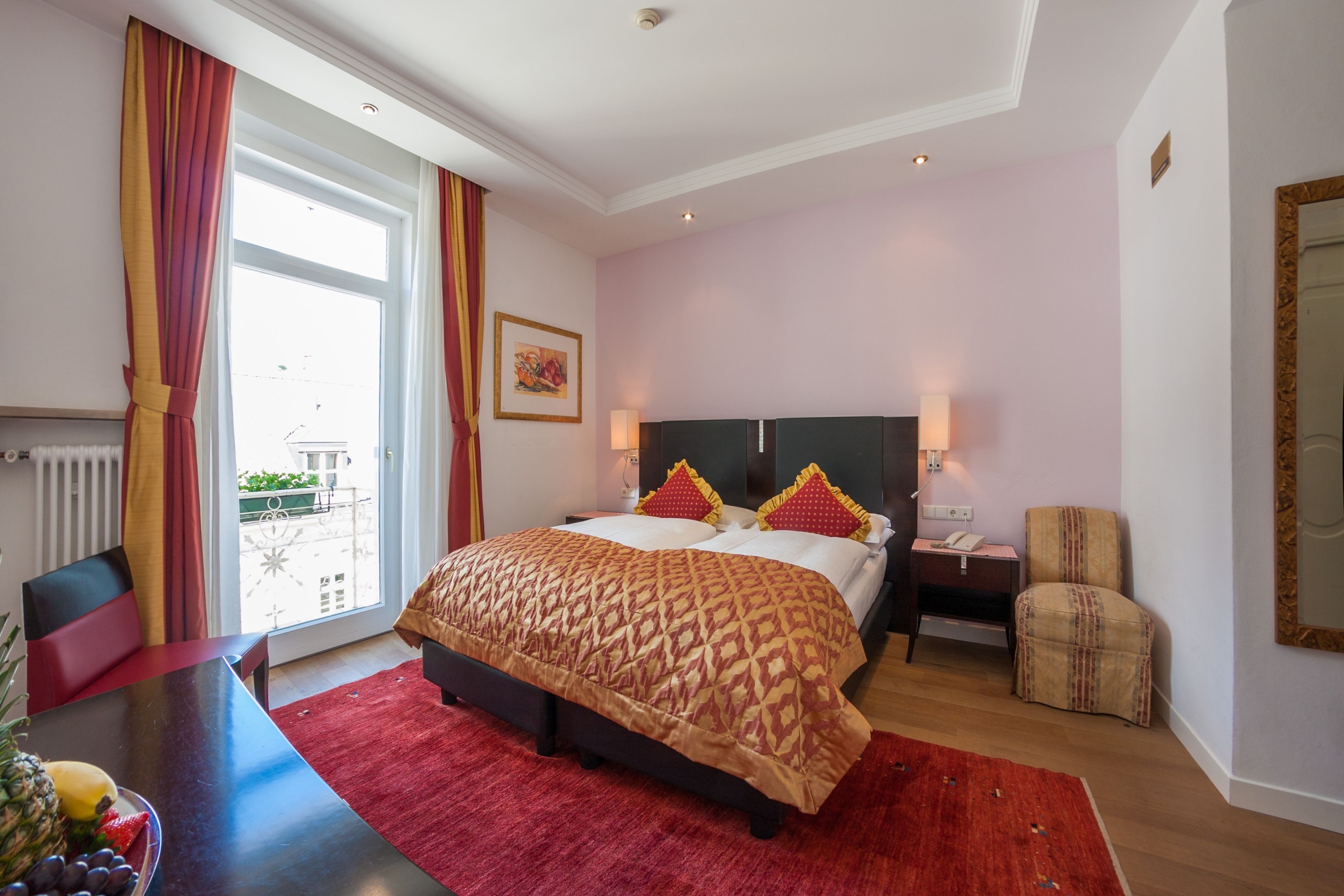 Ein Hotelzimmer mit einem großen Bett, einem Esstisch mit Früchten, einem Stuhl, einem Nachttisch und Blick ins Freie.