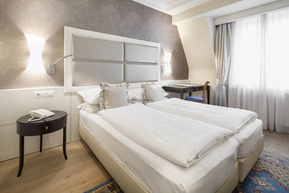 A hotel room with a large bed, a bedside table with a phone, a desk, and a window with curtains.