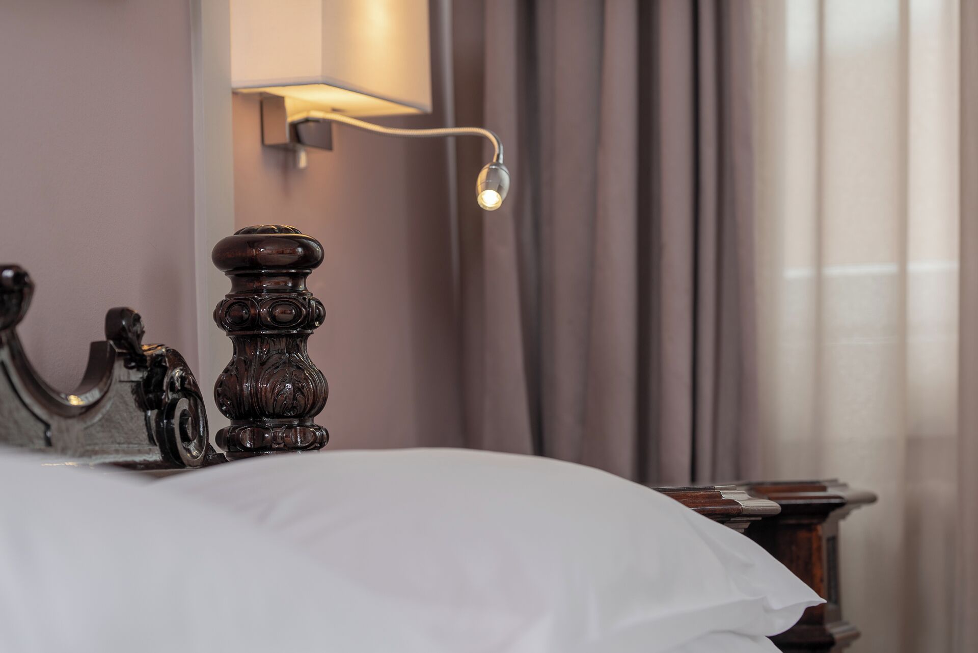 Ein Bett mit weißer Bettdecke, ein Holzkopfende mit aufwendigen Schnitzereien, eine Wandleuchte und ein Fenster mit Vorhängen.