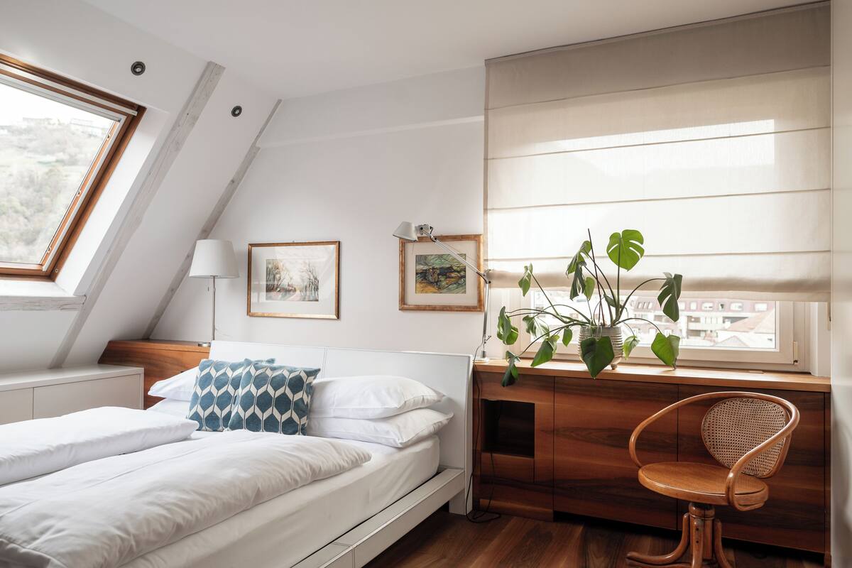 A bedroom with a bed, bedside table, lamp, framed pictures, a window with blinds, and a potted plant.