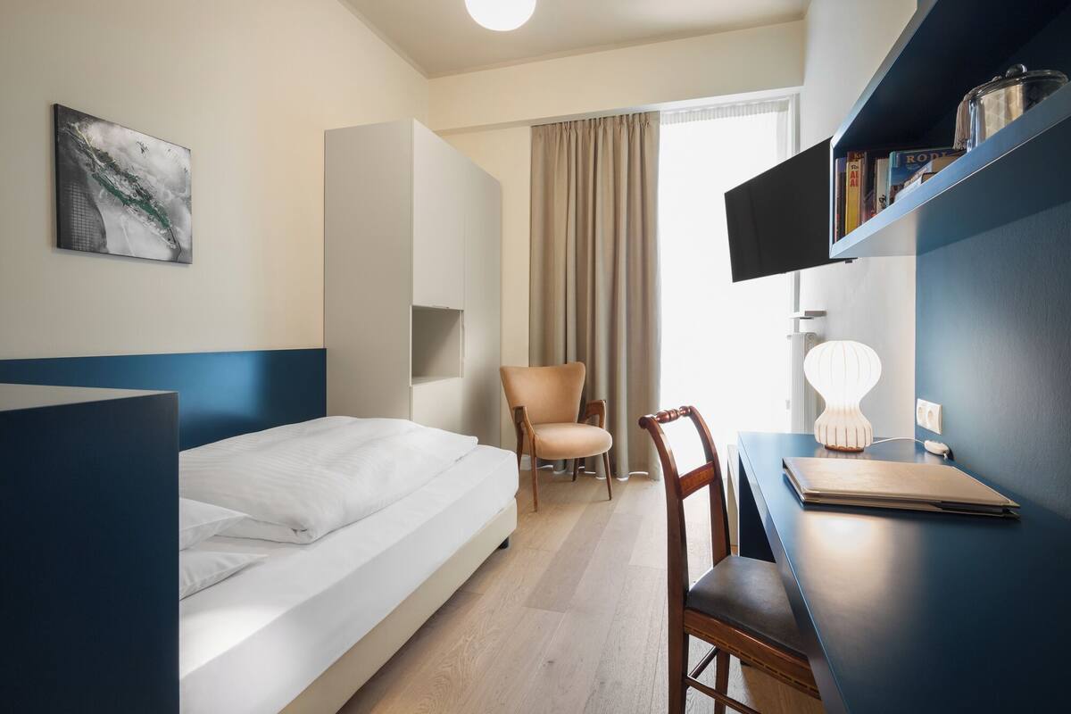 A hotel room with a bed, a desk, a chair, a television, and a bookshelf.