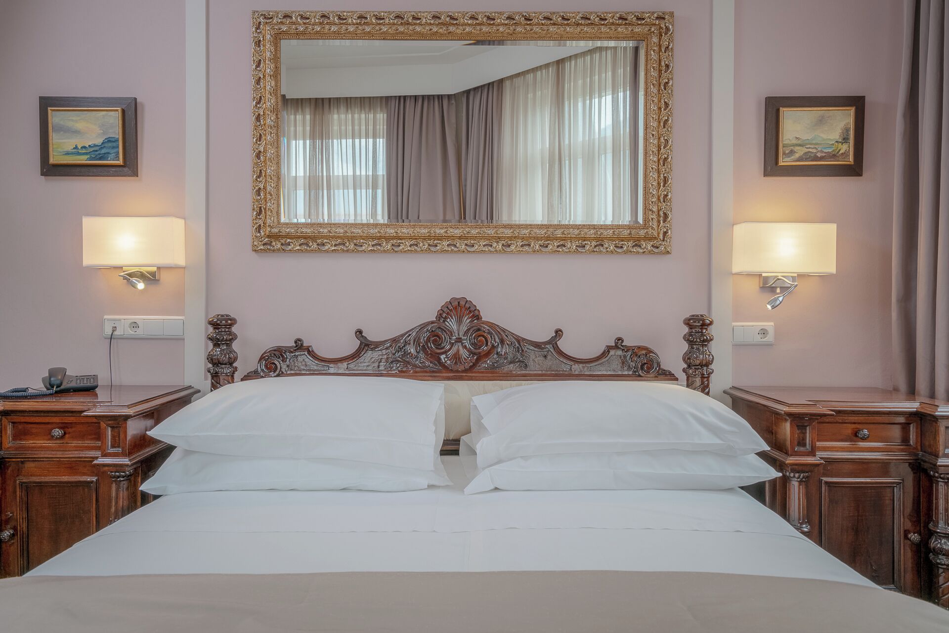 A hotel room with a large bed, ornate headboard, two bedside tables with lamps, a mirror above the bed, and two framed pictures on the walls.