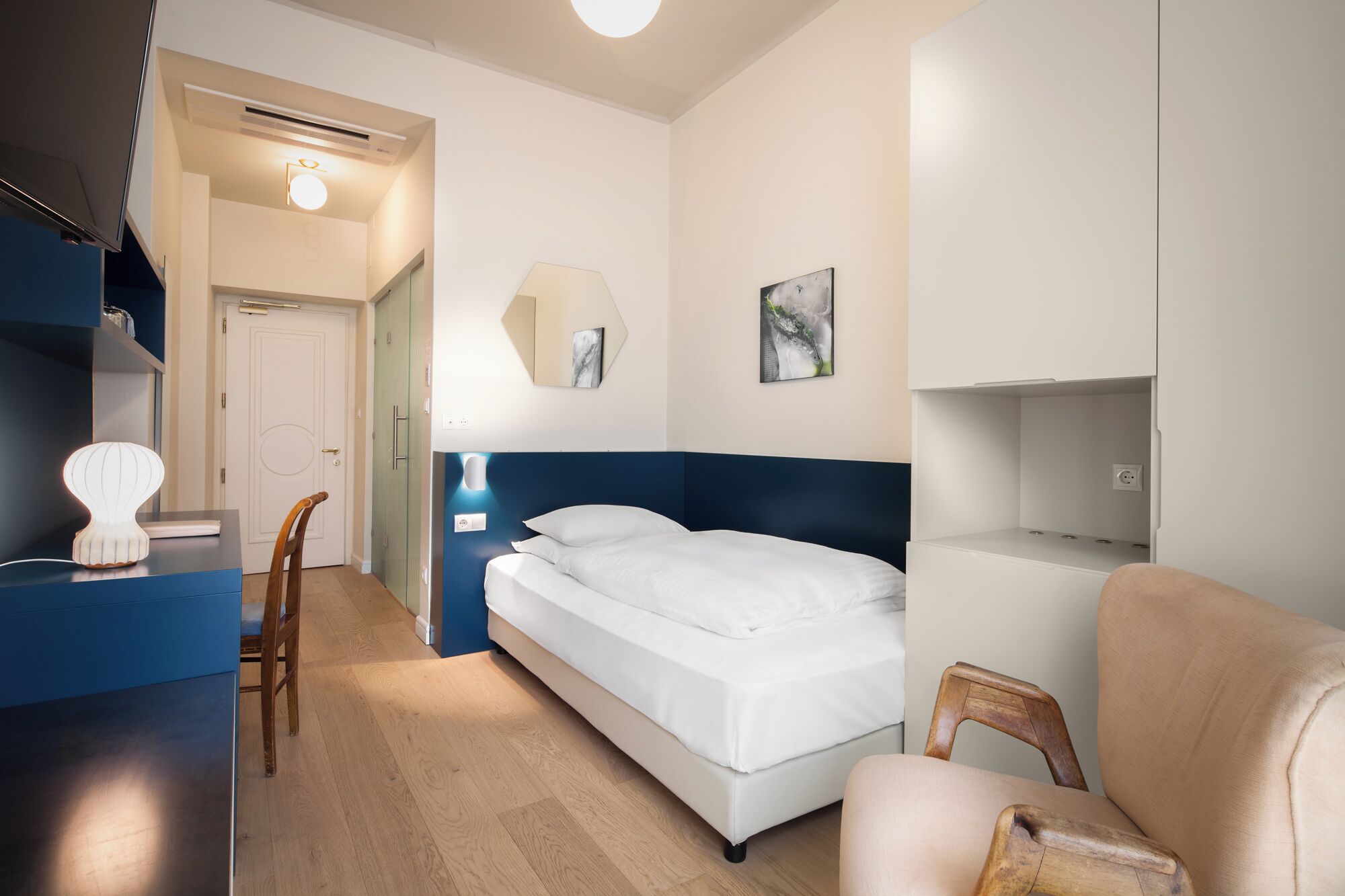 Un dormitorio con cama, una silla, un escritorio, una lámpara, un estante en la pared y una puerta.