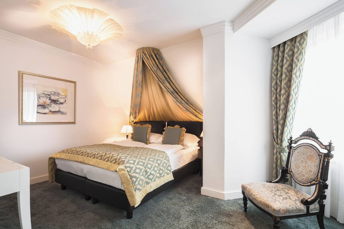 A bedroom with a bed, a chair, a desk, a painting, and a canopy.