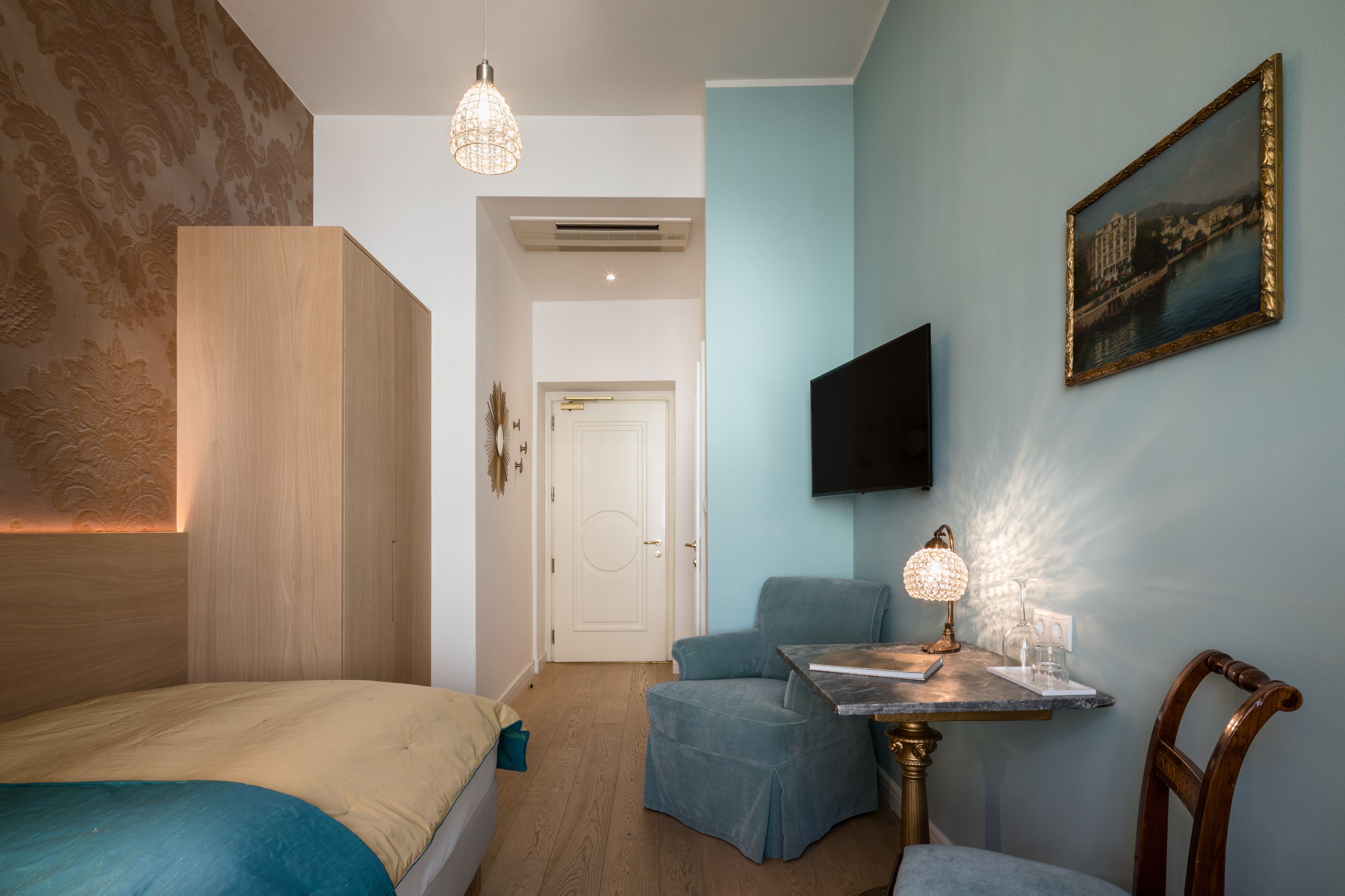 A bedroom with a bed, a desk with a lamp, a chair, a TV mounted on the wall, and a door leading to another room.