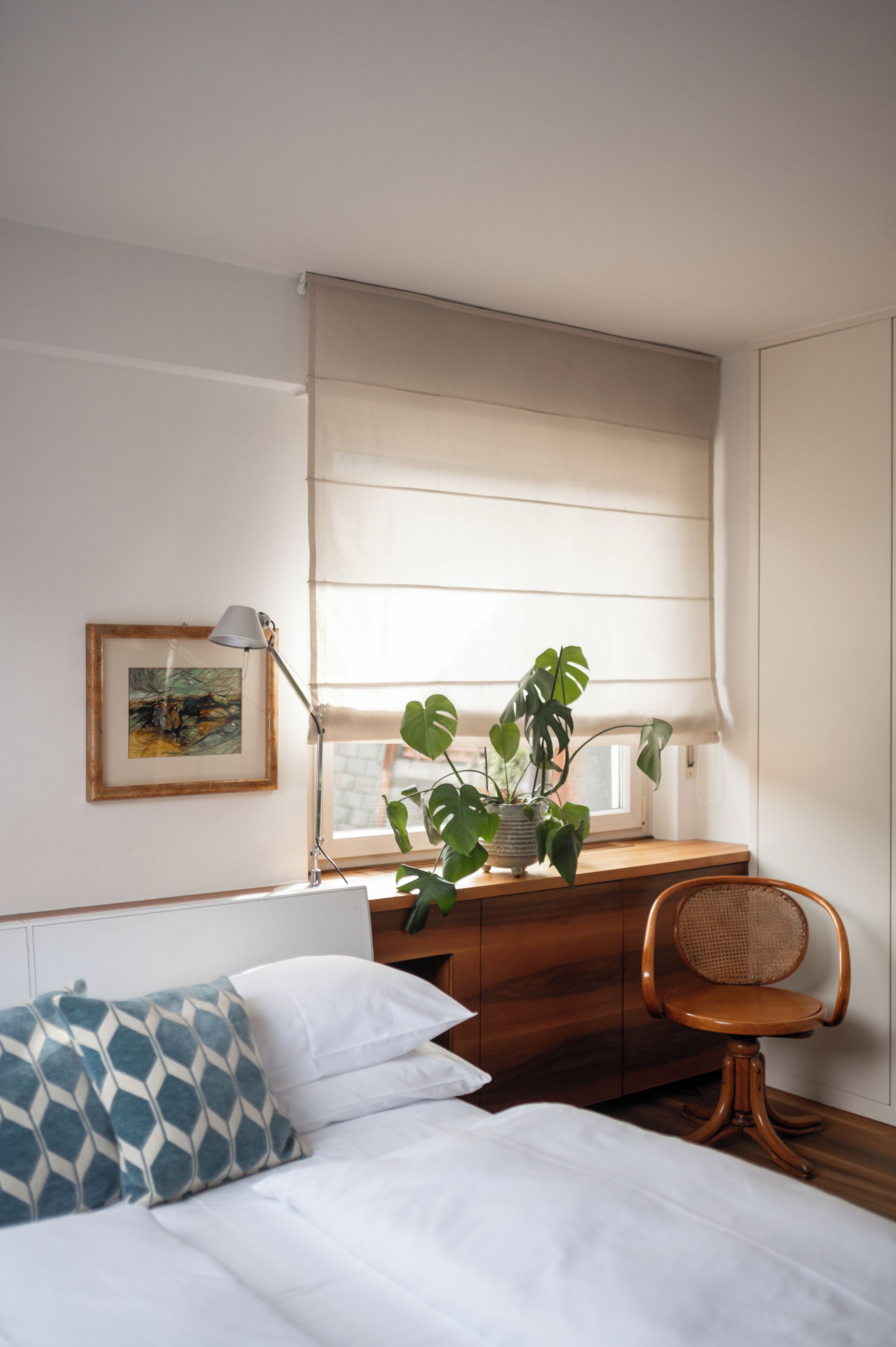 A bedroom with a bed, a desk with a plant, a chair, a lamp, and a window with blinds.