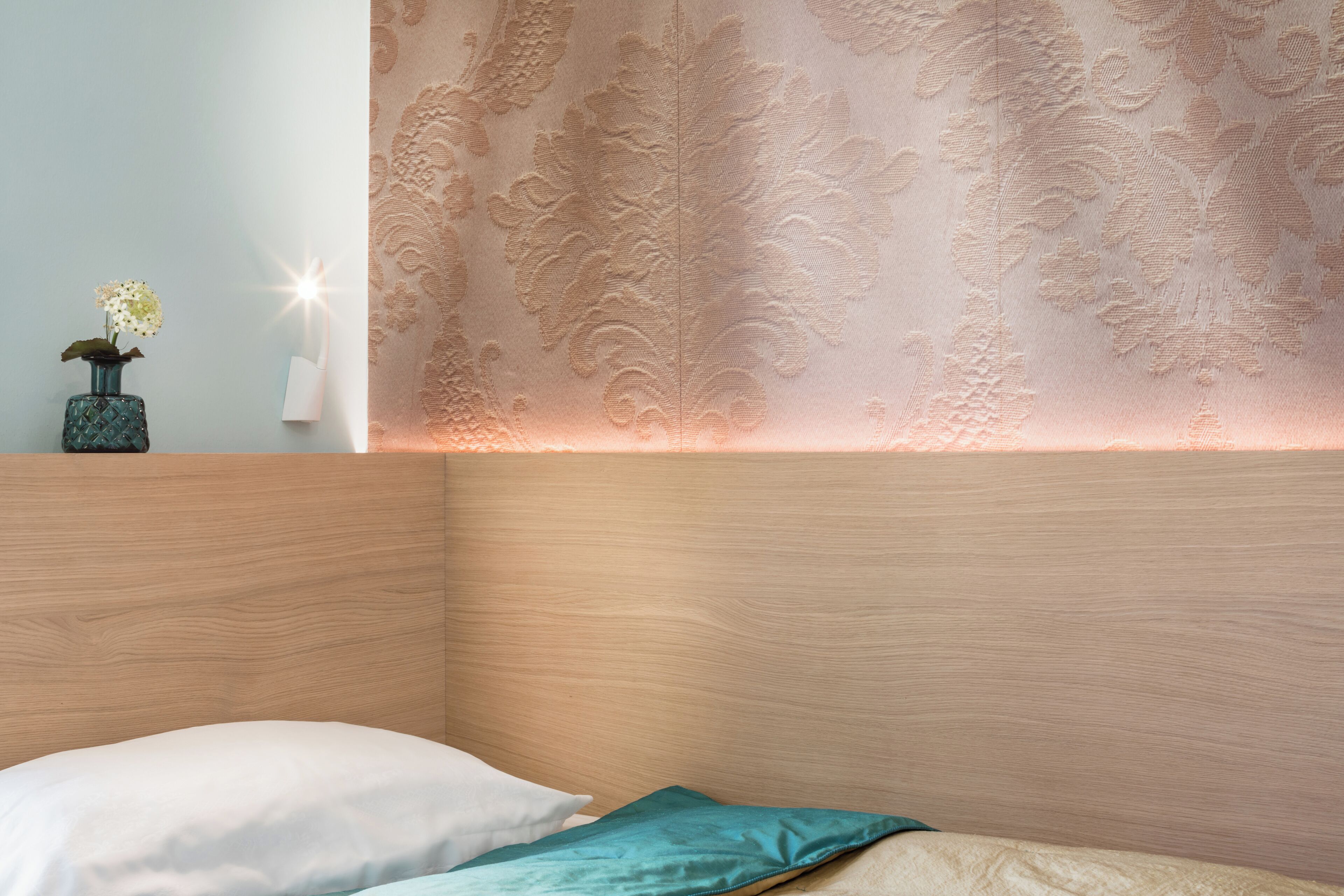A bedroom with a wooden headboard, a white pillow, a teal pillow, and a wall with a floral pattern.