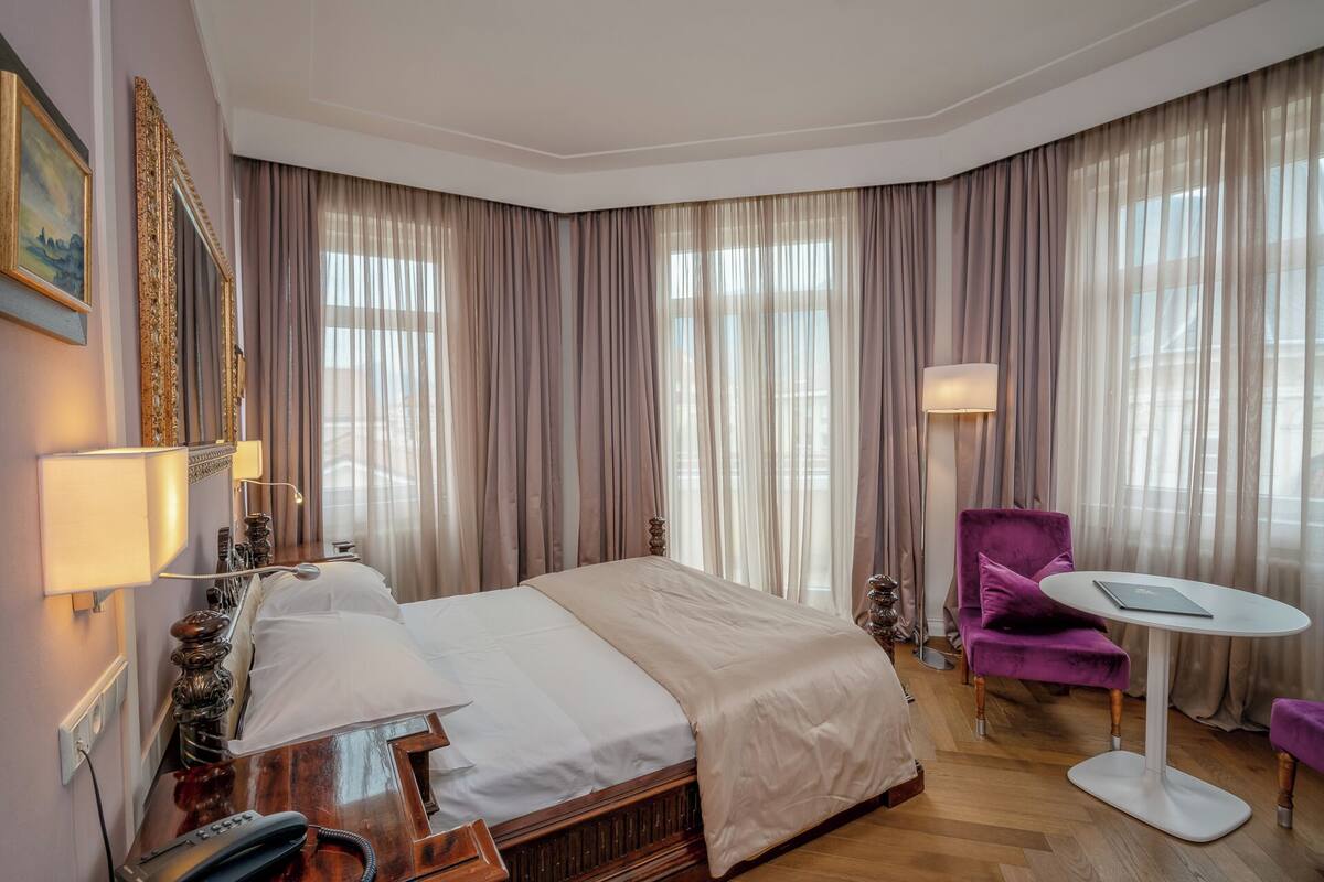 A hotel room with a bed, a desk, a chair, a lamp, and a large window with curtains.