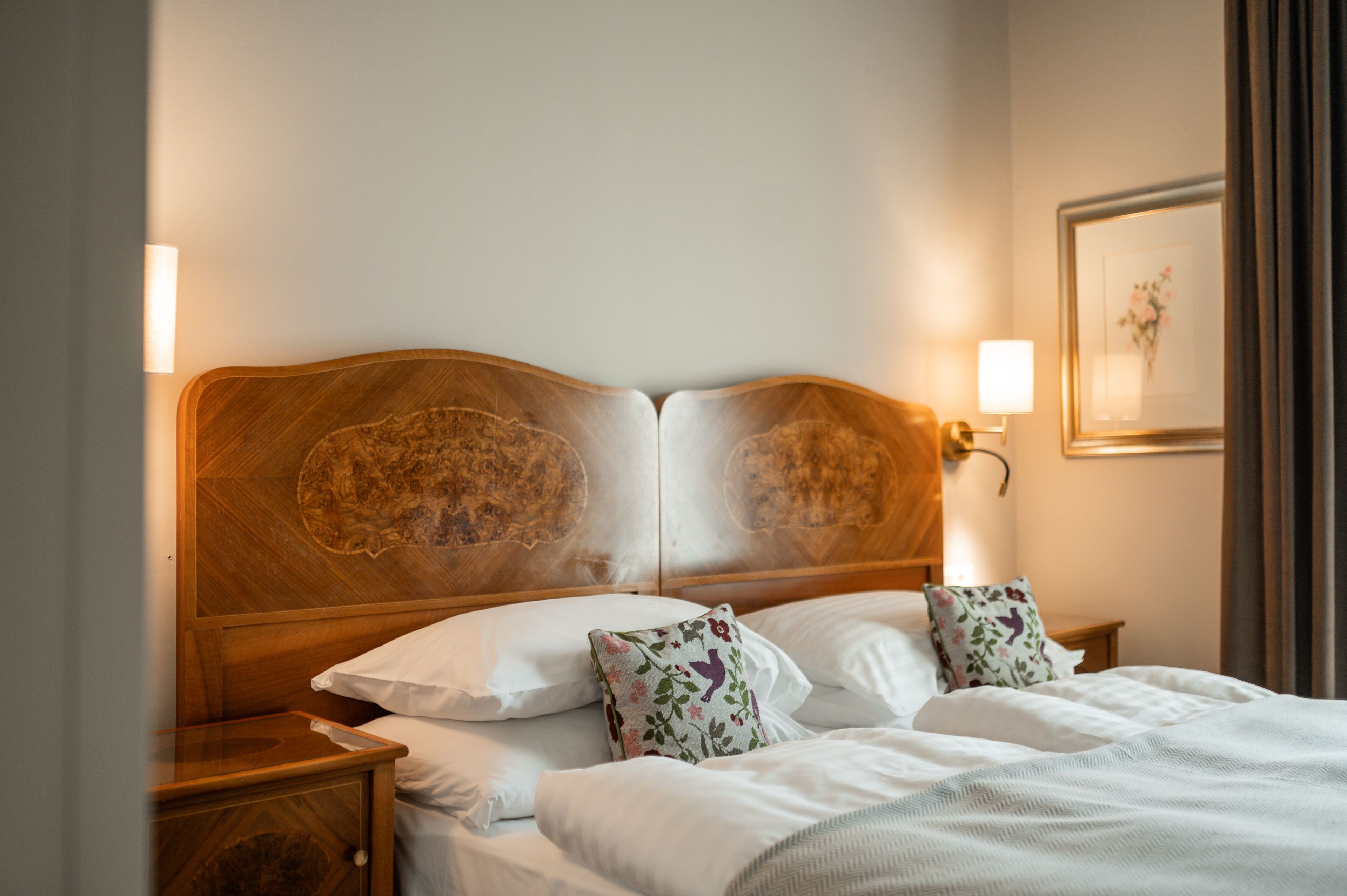 Ein ordentlich bezogenes Bett mit weißen Leinentüchern, floralen Kissen, einem Holz-Kopfschild und Nachttischlampen.