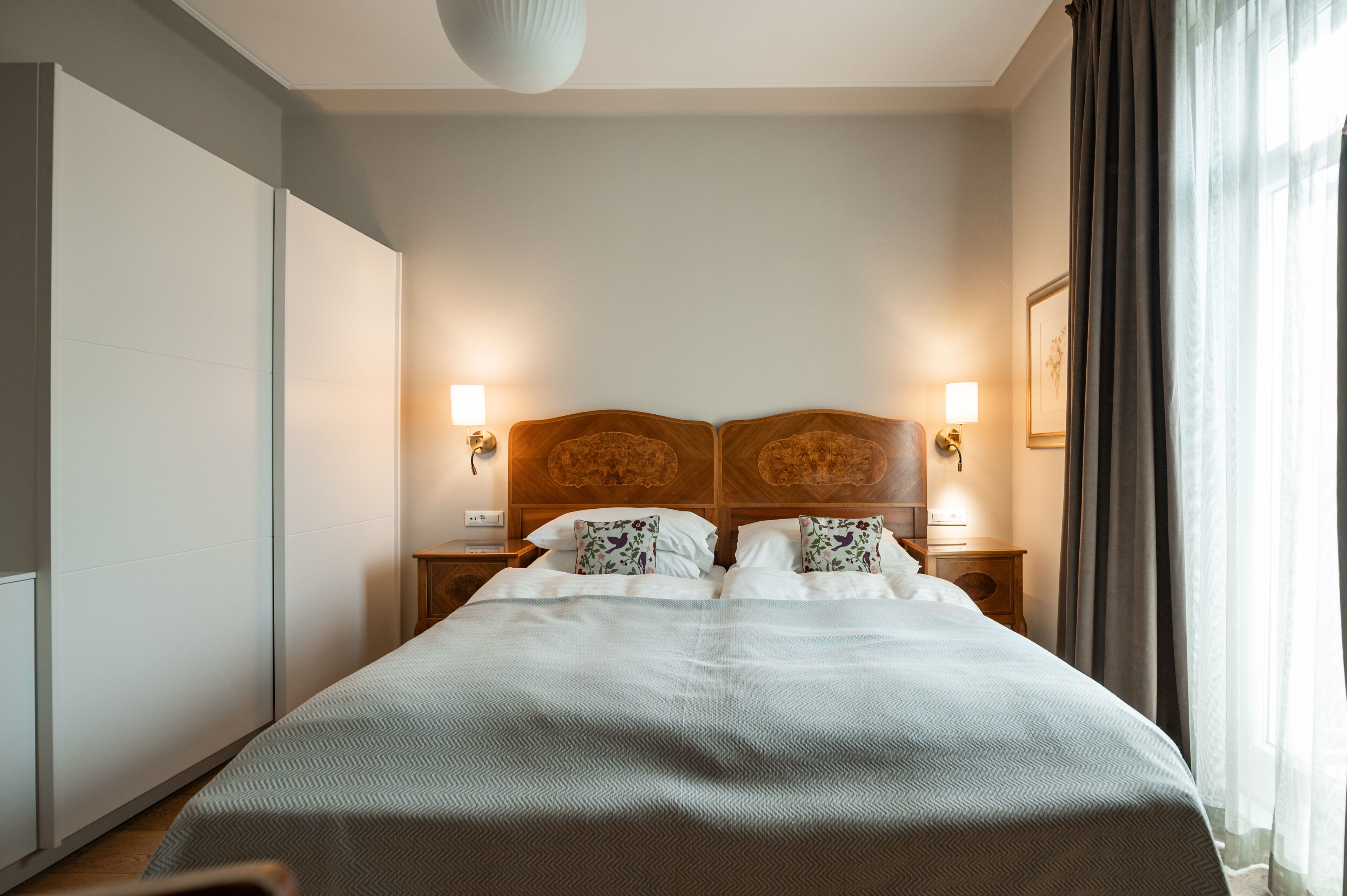 A neatly made bed with a wooden headboard, two bedside tables with lamps, and a large window with curtains.