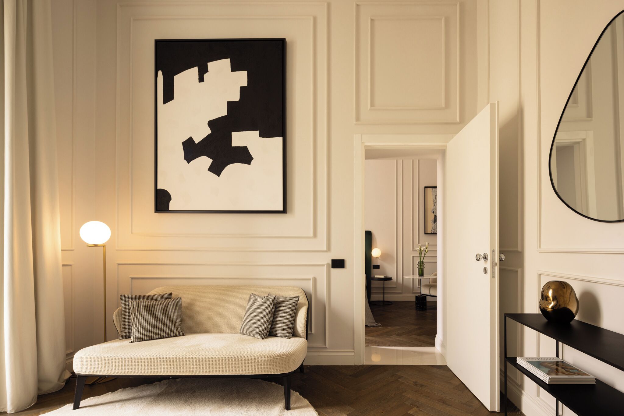 A modern interior with a sofa, a large abstract painting, a round mirror, and a hallway leading to another room.