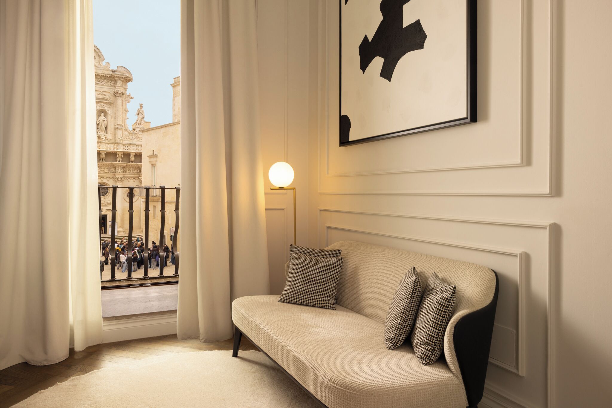 A room with a sofa, a framed artwork, a lamp, and a view of a building through a window.