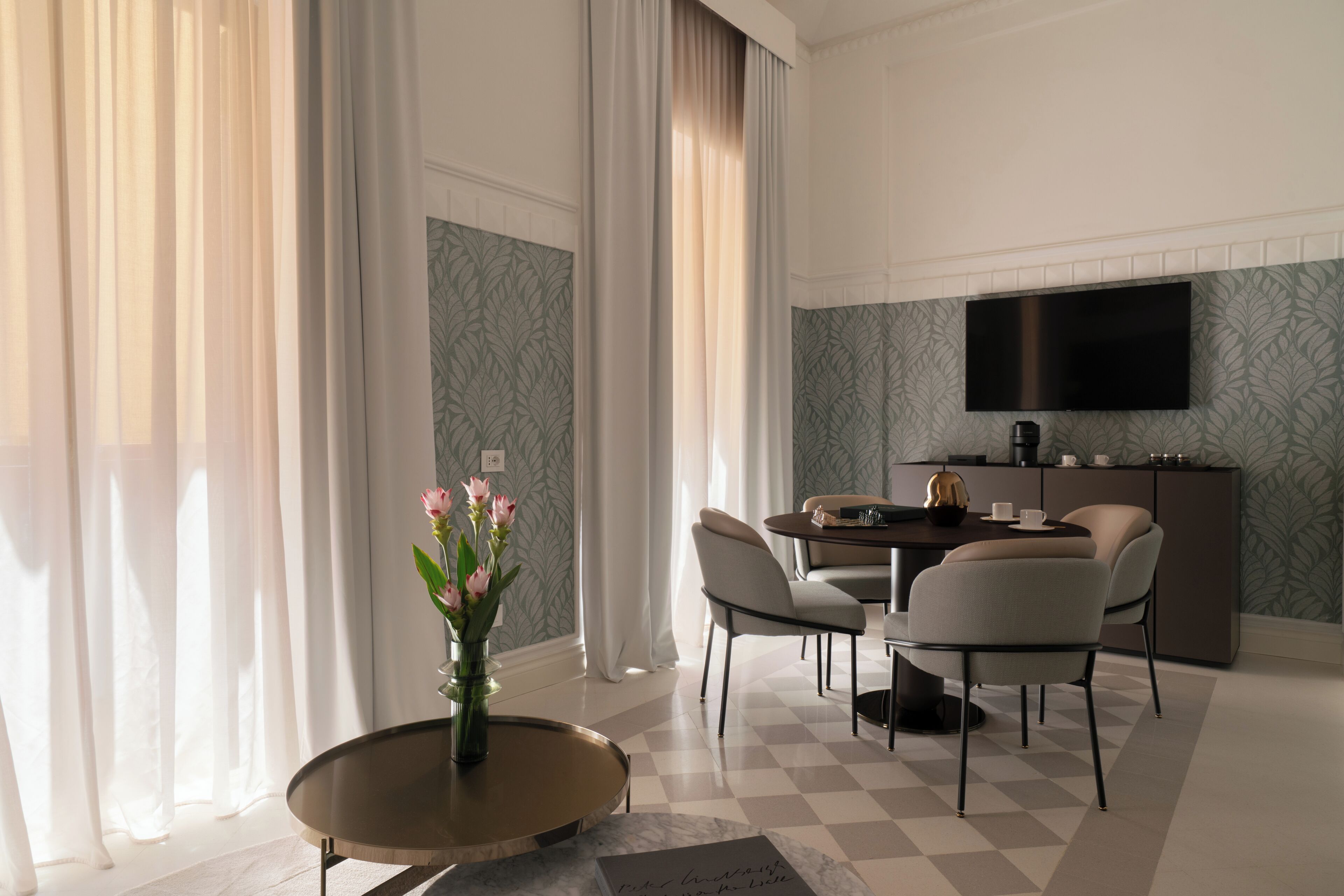 A modern hotel room with a dining area, a flat-screen TV mounted on the wall, and a vase with flowers on a round table.