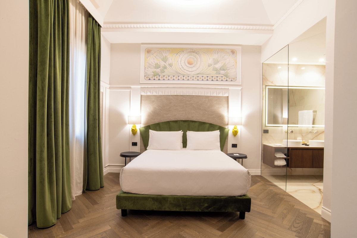 Quarto de hotel com uma cama grande, cortinas verdes, chão de madeira, banheiro moderno e um objeto decorativo sobre a cama.
