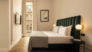 A hotel room with a bed, a nightstand, a framed picture, a window with a view of a building, and a door leading to an outdoor area.
