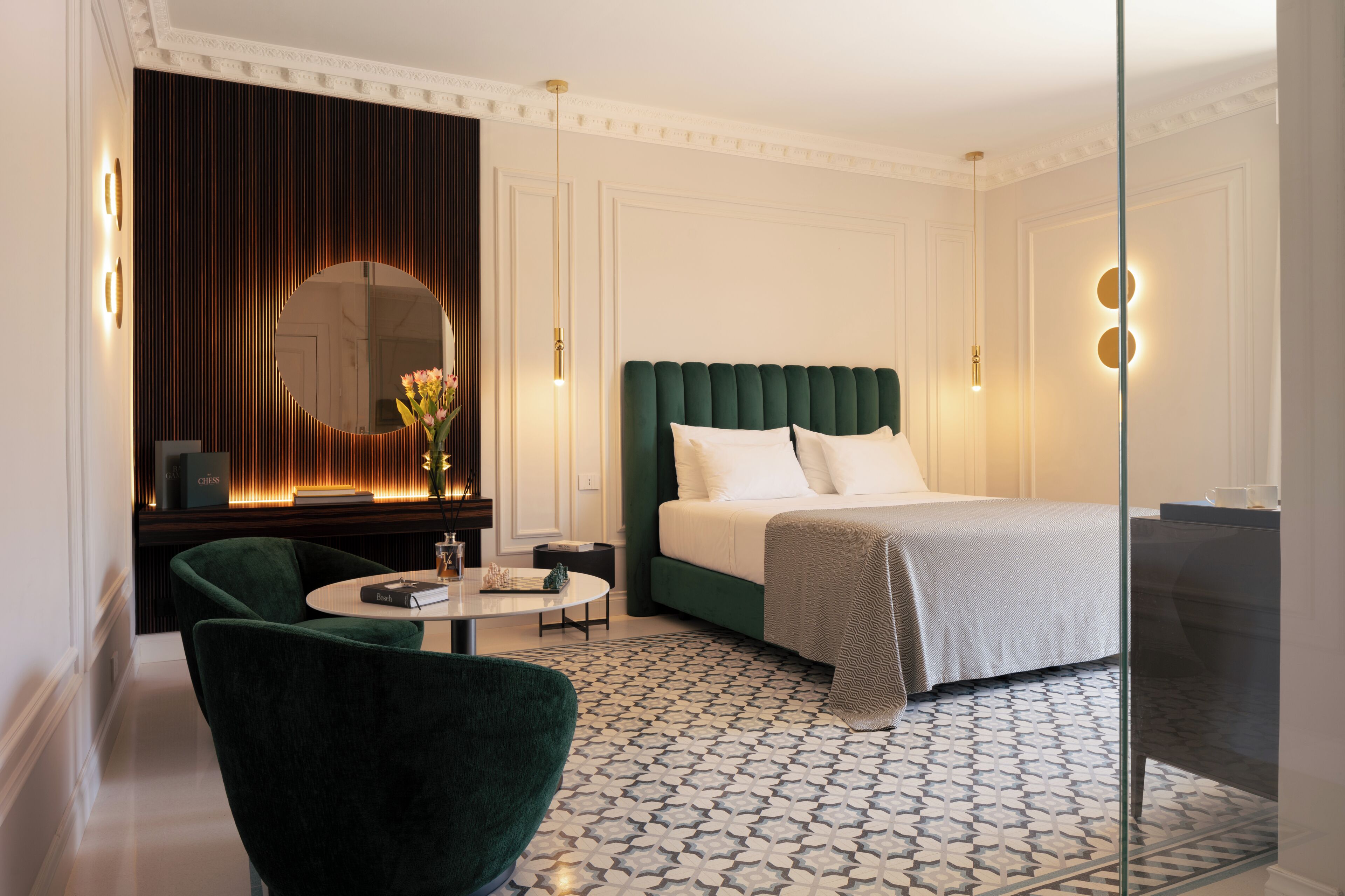 Hotelkamer met een bed, een ronde spiegel, een groene fauteuil, een klein tafeltje en een vloer met een patroon.