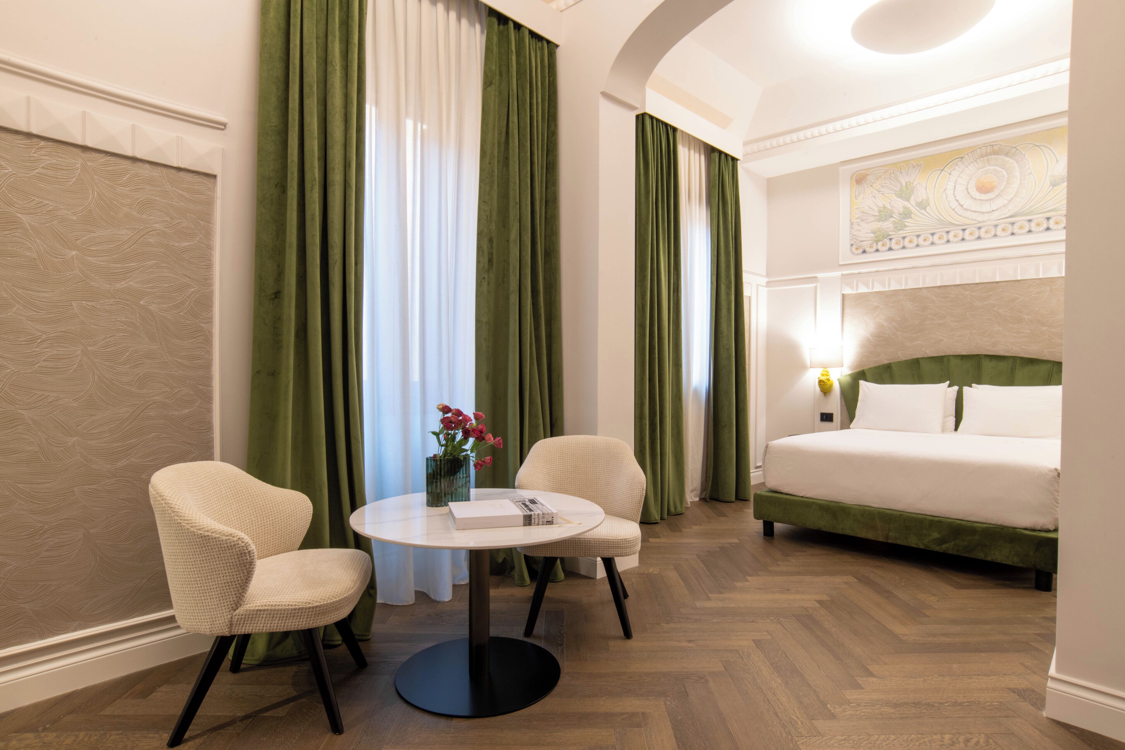 Una habitación de hotel con una cama, dos sillas, una mesa redonda y un florero con flores.