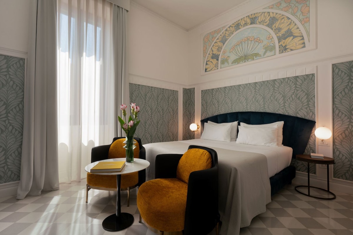 A hotel room with a bed, two chairs, a small table, a vase with flowers, and a decorative ceiling mural.