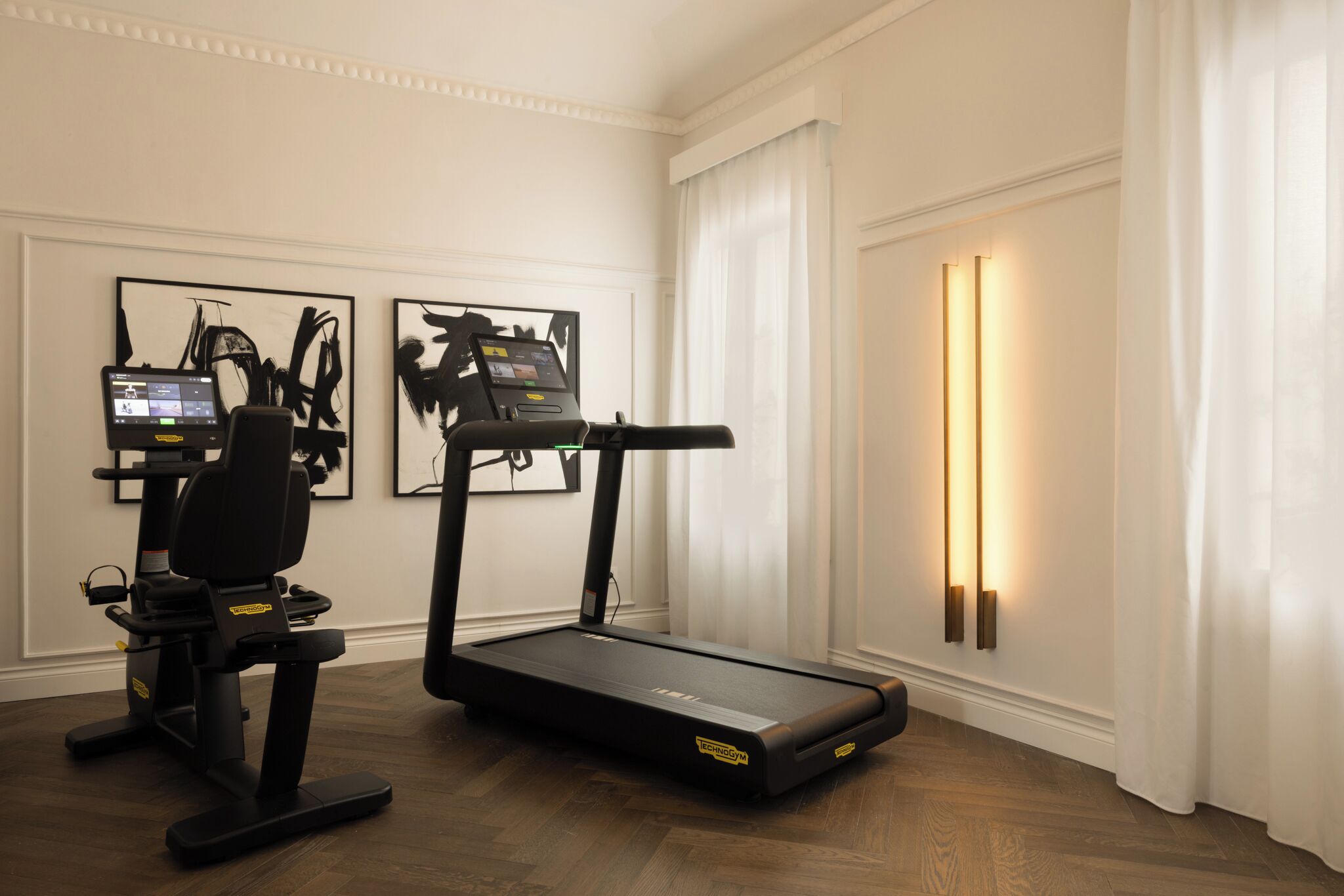 A treadmill in a room with white walls, a wooden floor, and two framed abstract paintings.