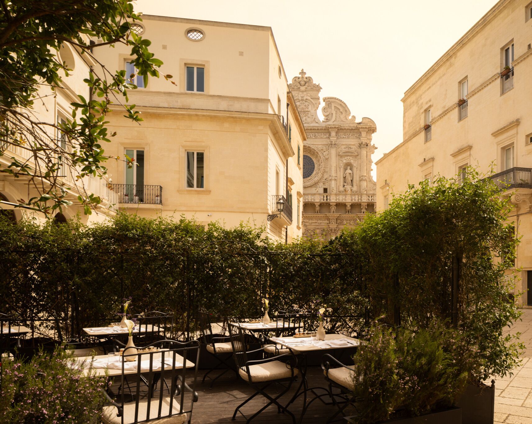 An outdoor café with tables and chairs, surrounded by greenery and historic buildings with ornate architecture.
