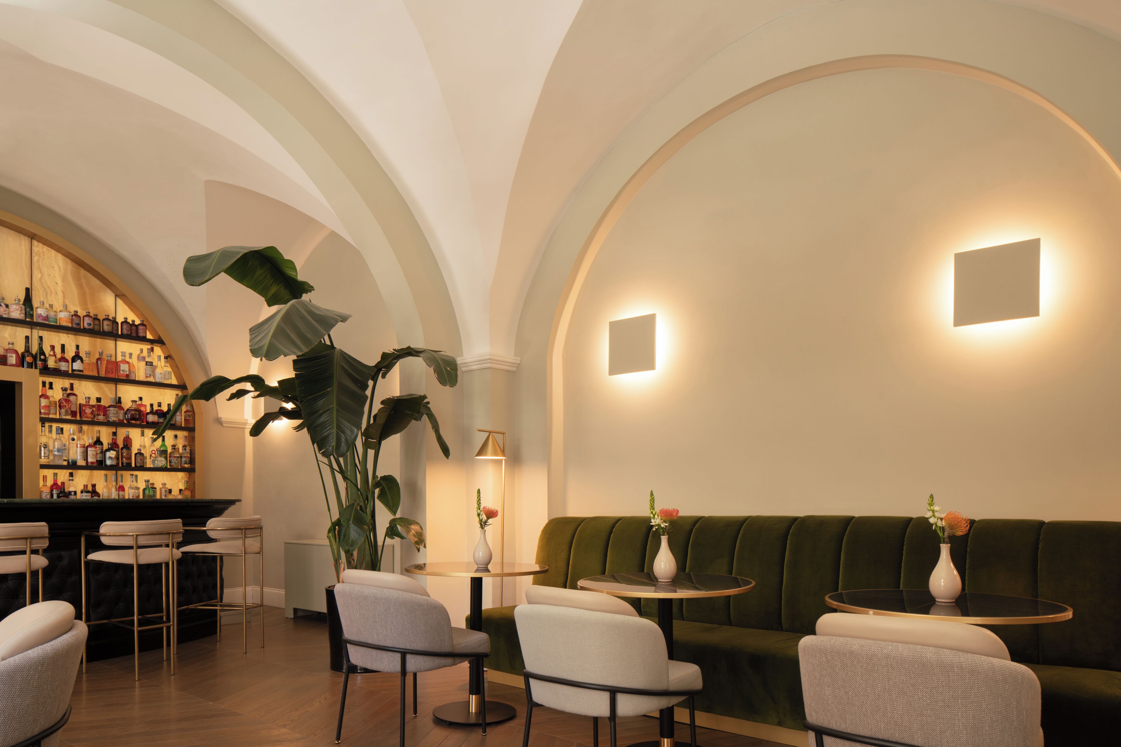 A bar area with shelves of bottles, a seating area with a green sofa and round tables, and a large plant.