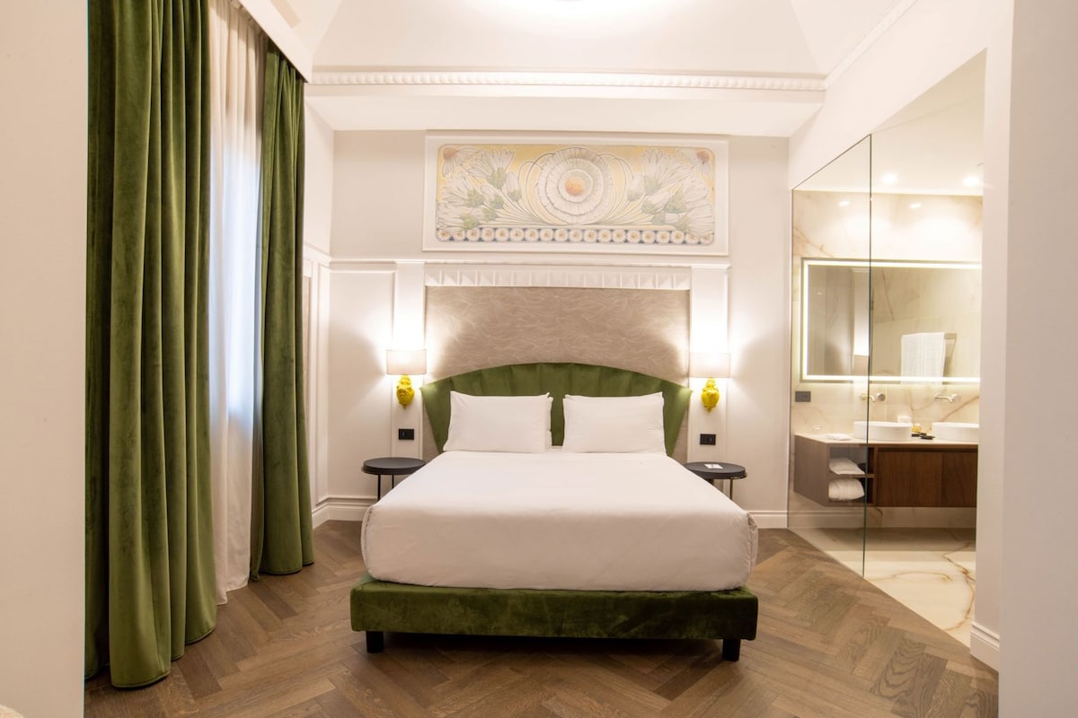 A hotel room with a large bed, a green headboard, a wall-mounted artwork, a bedside lamp, and a modern bathroom with a glass shower enclosure.