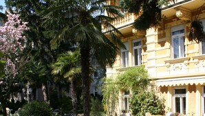 A courtyard with a palm tree, a garden with various plants, and a building with a balcony. - Hotel Westend (Merano)