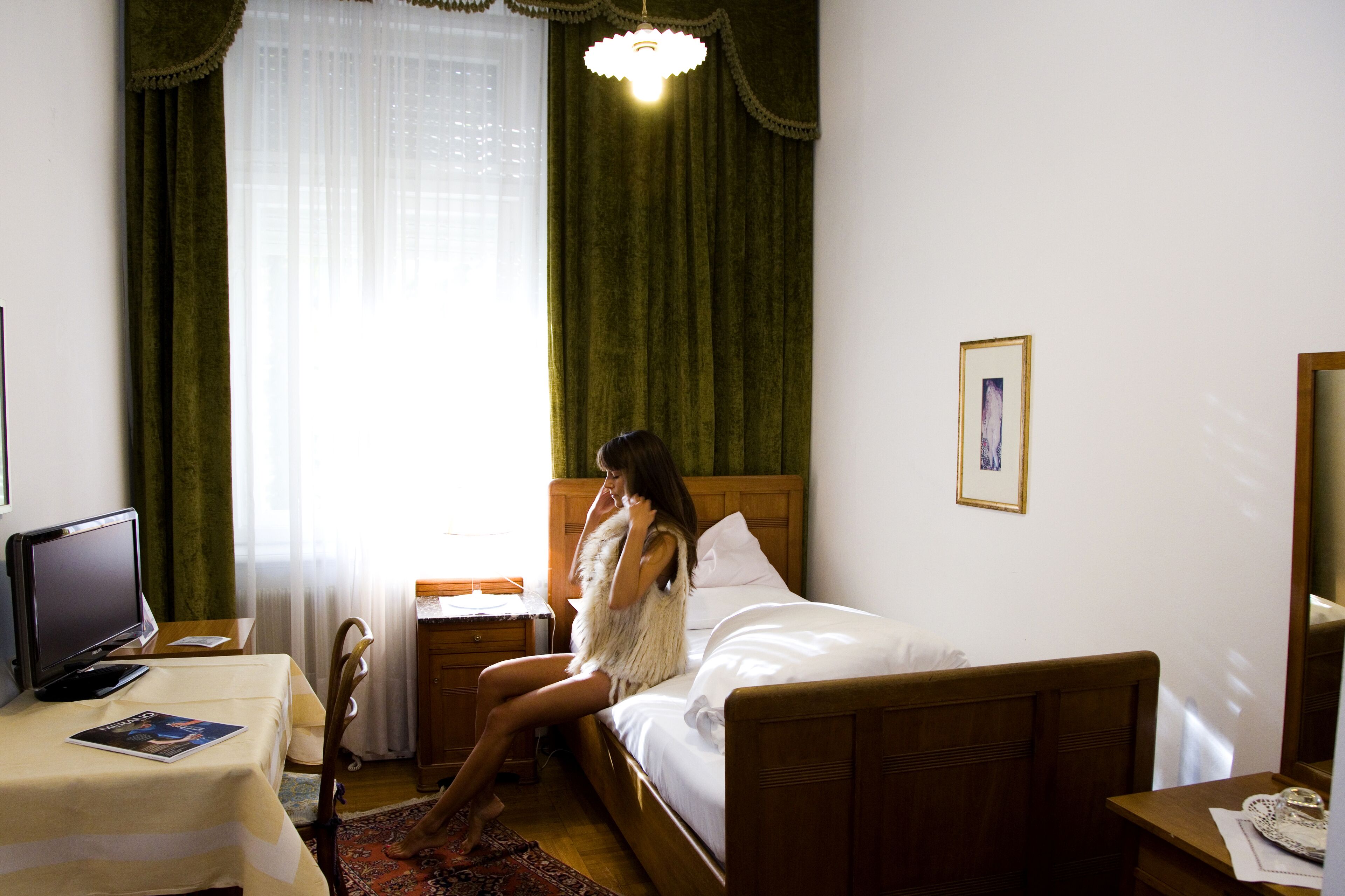 A hotel room with a bed, a desk with a computer, a chair, a small table, and a window with curtains.