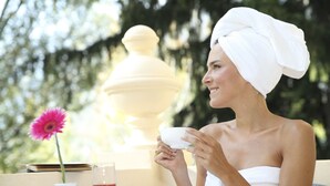 A person with a towel wrapped around their head, holding a cup, seated at an outdoor table with a vase of flowers, a candle, and a glass of red drink. - Hotel Westend (Merano)