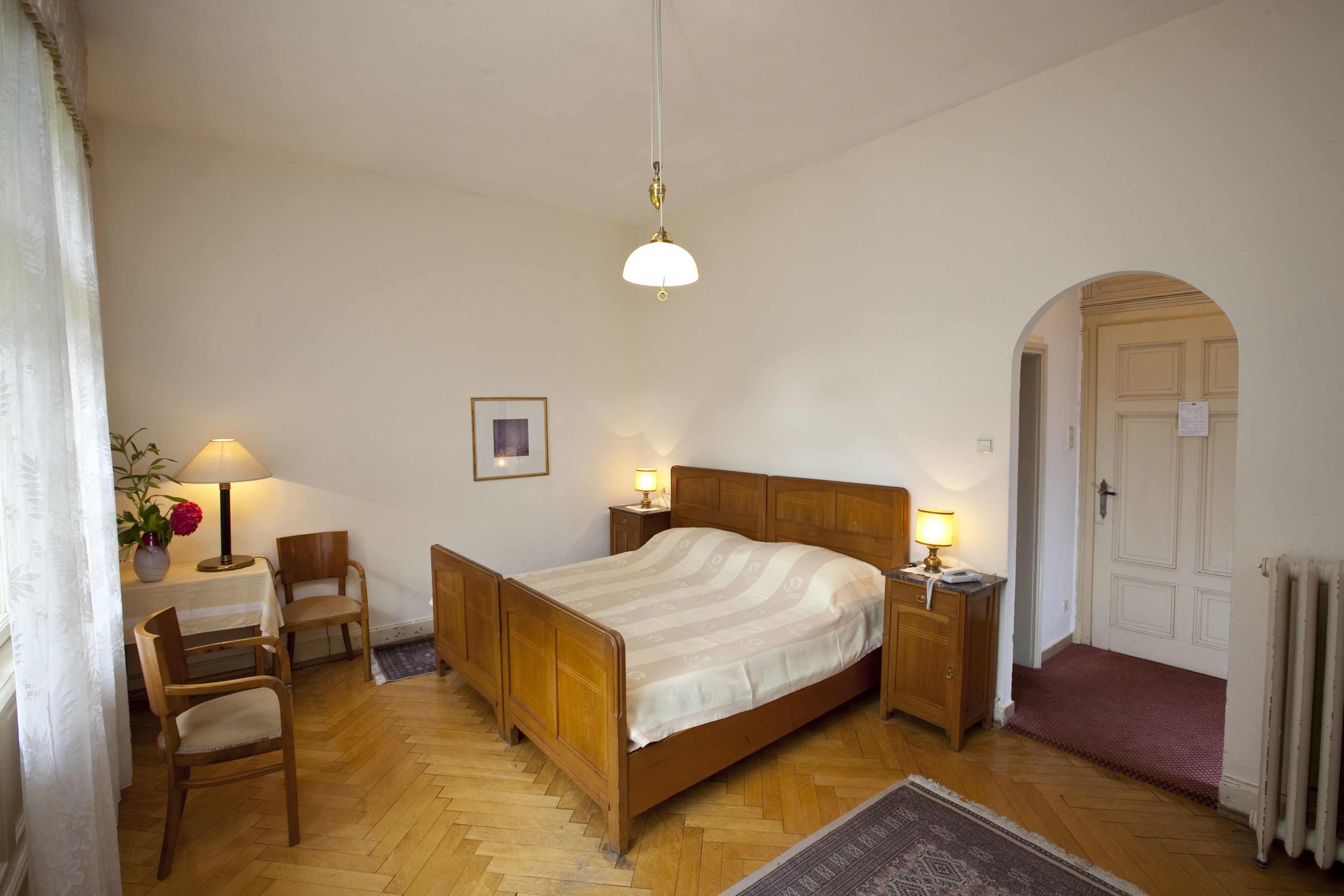 Une chambre avec un lit en bois, des tables de chevet avec des lampes, une chaise, un bureau avec une lampe, une plante en pot et une porte avec un paillasson.