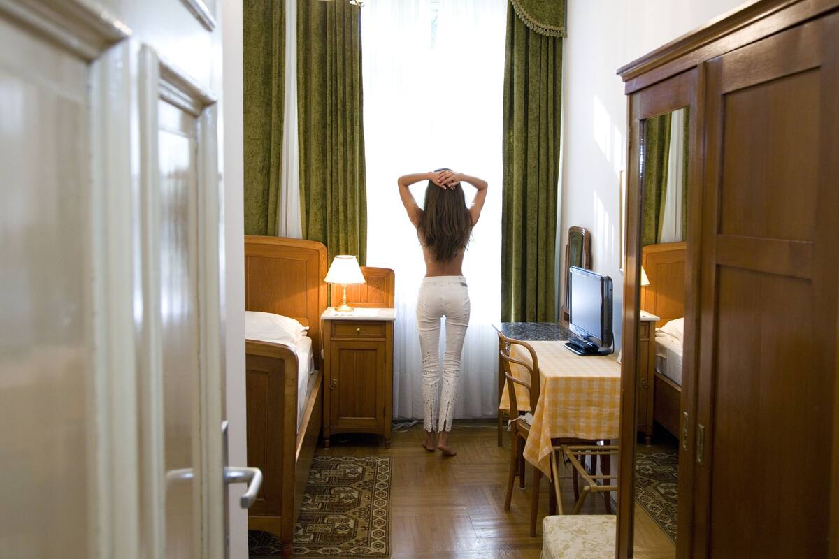 A hotel room with a bed, a desk with a computer, a chair, a lamp, and a window with curtains.