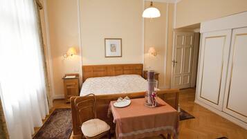 Una habitación de hotel con una cama, una mesa con mantel, una silla, una mesita de noche, una lámpara, un cuadro en la pared y un armario.