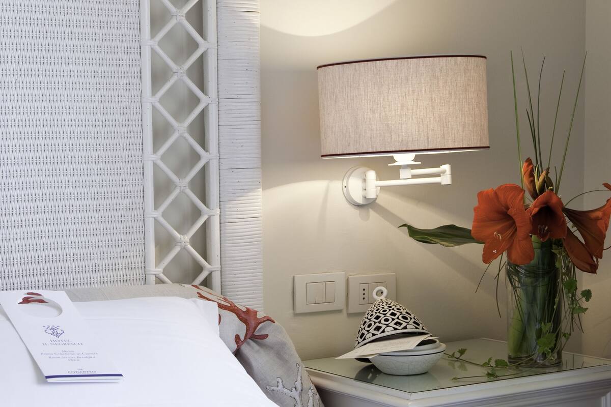 A neatly made bed with white linens, a bedside table with a vase of flowers, a lamp, and a decorative wall piece.